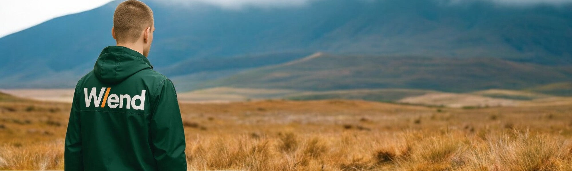 Person wearing a green hoodie with W/end logo, standing in a field with mountains in the background